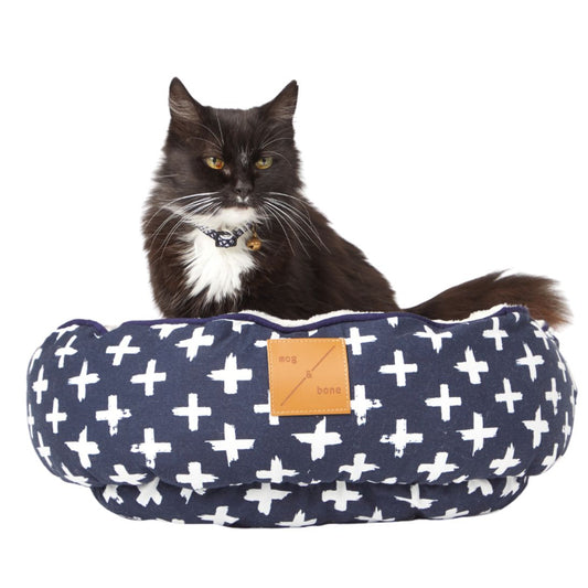Cat sitting in a navy blue pet bed with white cross patterns on a white background