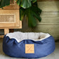 Blue cat bed with a brand label in a home setting with a plant and wooden furniture.