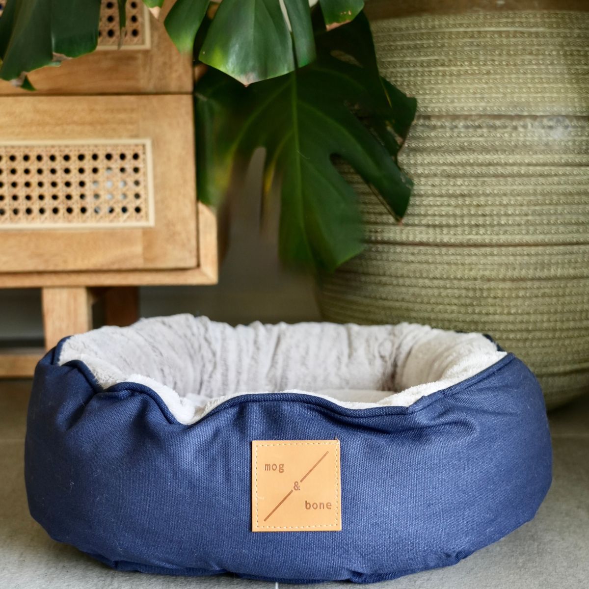 Blue cat bed with a brand label in a home setting with a plant and wooden furniture.